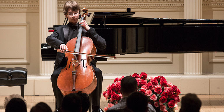 Andrew Bailey playing his cello in front of a grand piano in an ornate hall. Audience and roses in the foreground.