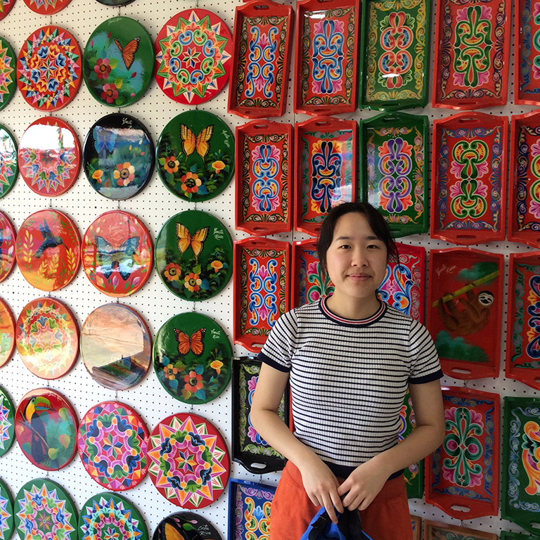 Ashley Bang stands in front of a wall covered in colorful painted art pieces.