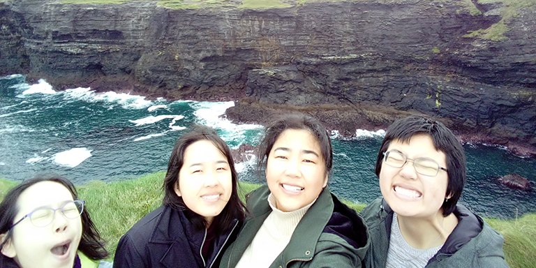 Ashley Bang and three friends smile near the ocean, cliffs in the background.